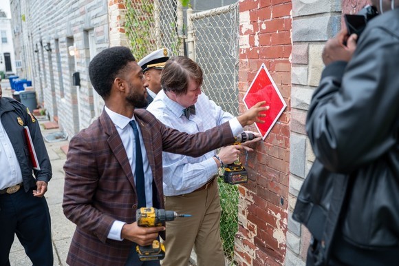 Photo of Mayor Brandon M. Scott and city employees mounting a red diamond shaped sign on a building